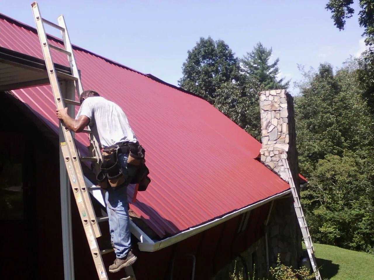Roofer on a ladder beside a red metal roof and stone chimney — Soffit & Fascia Repair by Roofing Brothers in Slovan